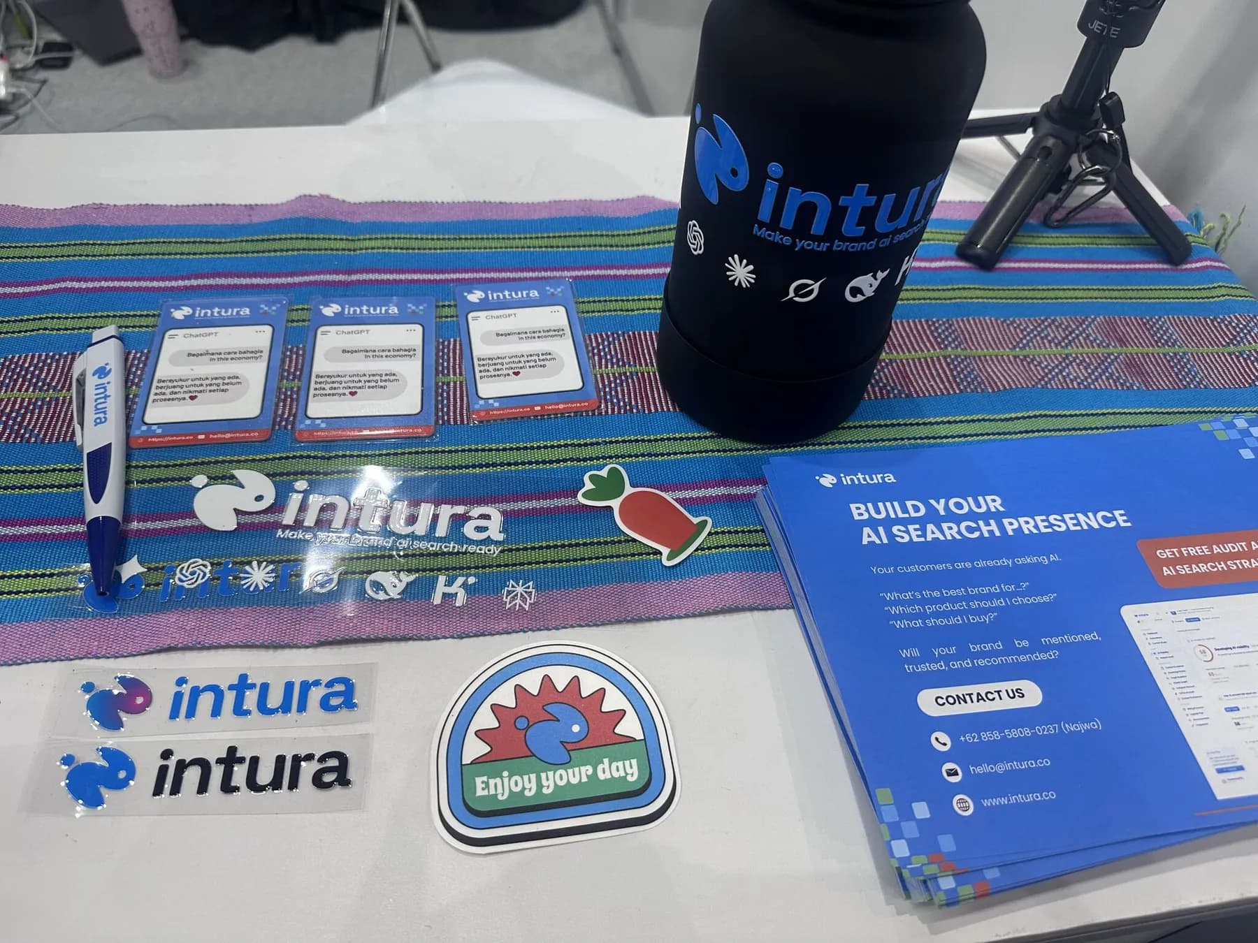Intura booth giveaways at INTI 2026 — stickers, pens, a branded tumbler, and flyers headlined "Build your AI search presence"