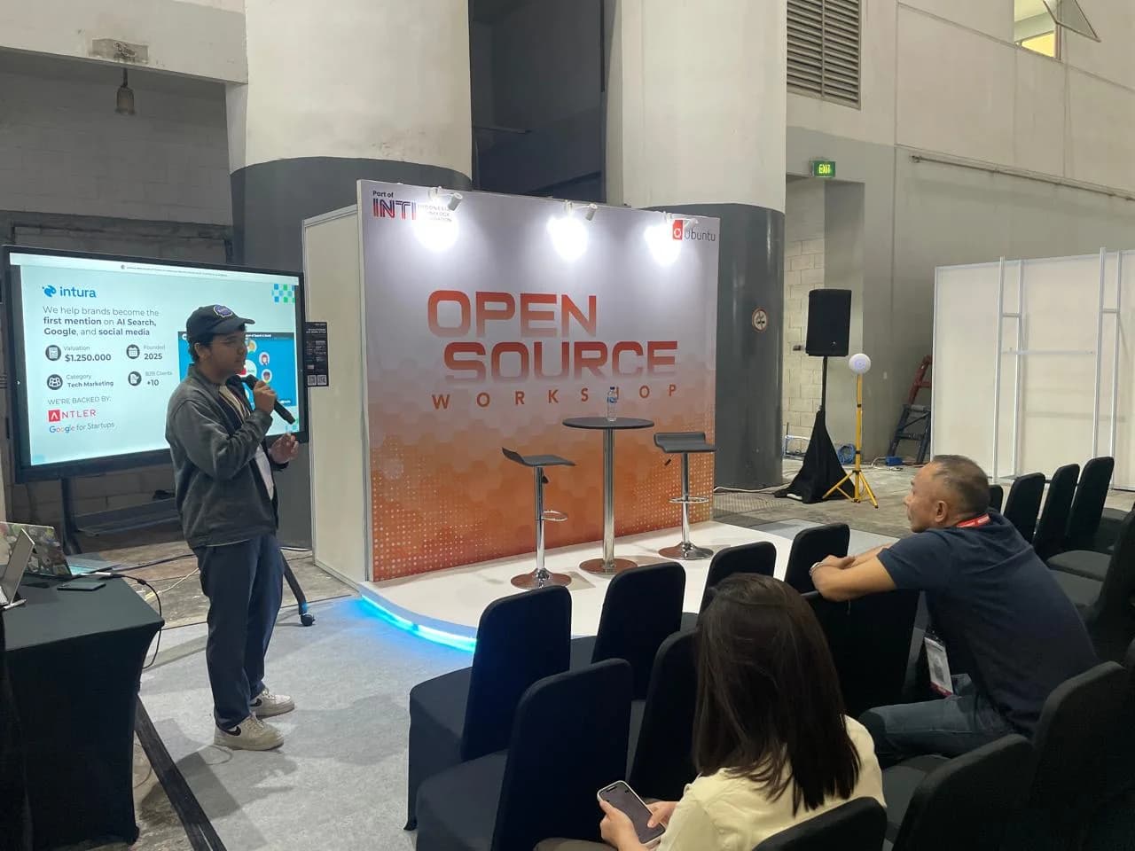 Intura presenting on the Open Source Workshop stage at INTI 2026 — the slide reads "We help brands become the first mention on AI Search, Google, and social media"